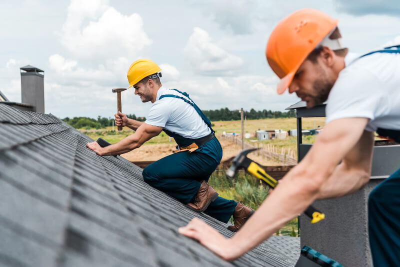 Workers on a residential roof using a hammer to make repairs to asphalt shingles.